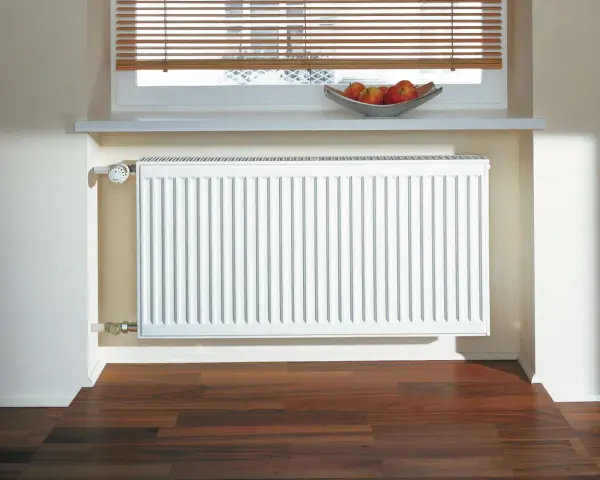 Radiador blanco de aerotermia con aletas de aluminio, bajo una ventana con persiana y un cuenco de manzanas.
