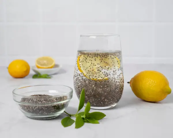 Vaso con agua de chia, rodaja de limón y semillas de chía en un bol. Limones y hojas verdes completan la escena.