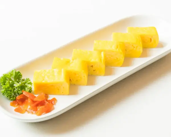 Sześć kawałków puszystego omletu tamago ułożonych na białym talerzu, z gałązką pietruszki i pomarańczowymi dodatkami.