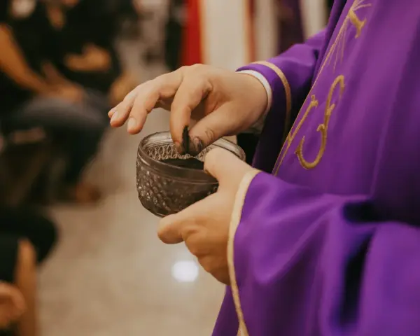 A priest in a purple robe takes ashes from a small bowl, preparing for the first day of Lent prayer.