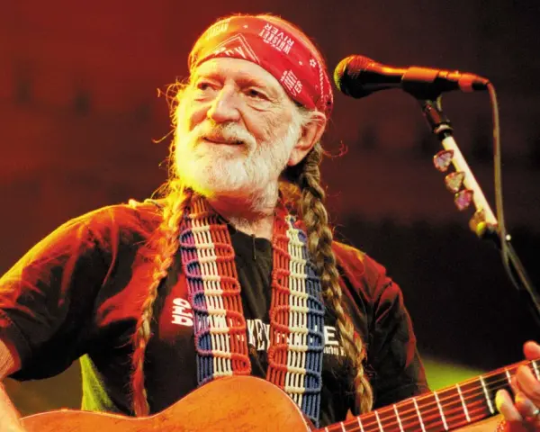 Willie Nelson mit roter Bandana und Zöpfen spielt Gitarre. Ein Mikrofon steht bereit für den Titel von Willie Nelson.