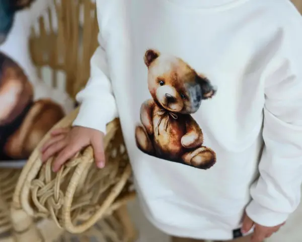 Biała bluza Moschino z uroczym misiem w stylu vintage. Dziecko trzyma się wiklinowego koszyka.