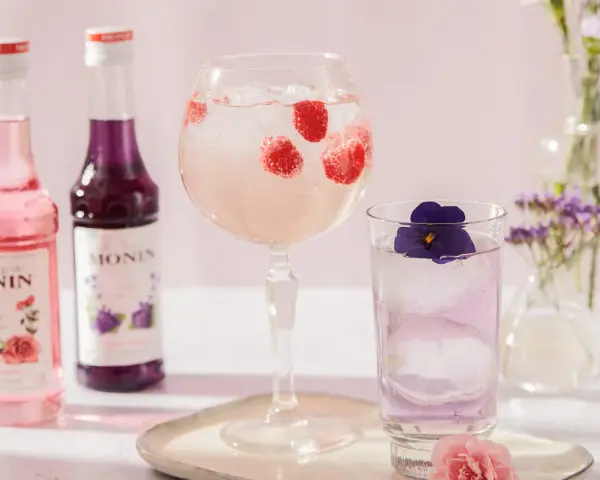 Dobre drinki przepisy: różowy i fioletowy syrop Monin, drink z malinami w kielichu, drink z fiołkiem w szklance, kwiaty w wazonach.