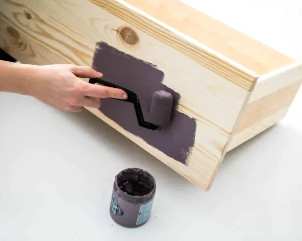 En hand använder en liten roller för att måla en trälåda med mörklila färg, vilket ger en jämn yta utan penseldrag.