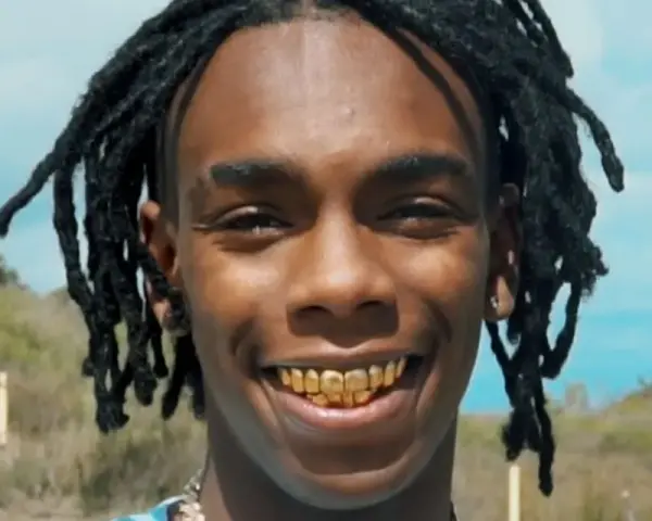 YNW Melly, uśmiechnięty z złotymi zębami, na tle nieba i wzgórz.