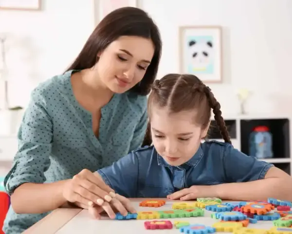 Nauczycielka wspiera dziewczynkę z niepełnosprawnością intelektualną w stopniu lekkim, układającą kolorowe puzzle.