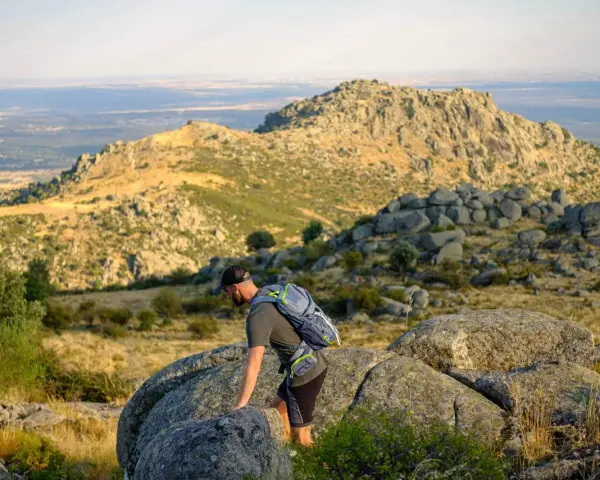 Un excursionista con mochila asciende por un terreno rocoso en la ruta Las Machotas, con un paisaje montañoso y un valle al fondo.