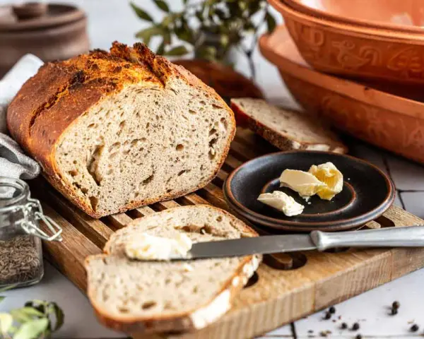 Ein rustikales Roggenbrot mit Sauerteig, aufgeschnitten und mit Butter bestrichen. Daneben ein Glas Kümmel und ein Messer.