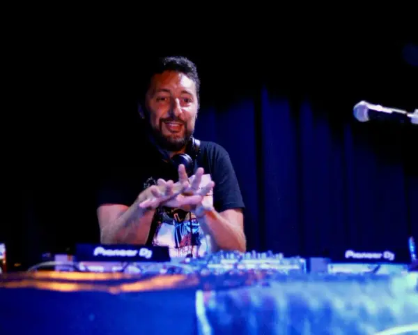 Un dj hip hop souriant, mains en l'air, mixe sur scène devant un public invisible.
