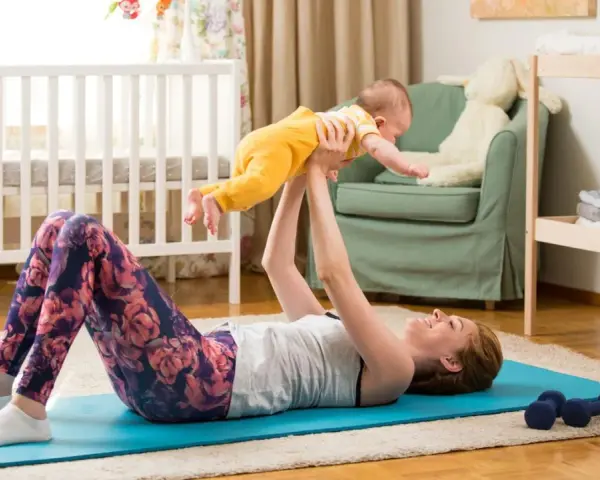 Frau macht Beckenbodentraining mit Baby. Sie liegt auf einer Matte und hebt ihr Kind hoch.