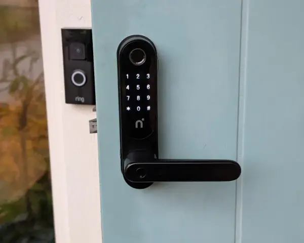 Nimly Touch Pro test: Svart digital dörrlås med kodpanel och fingeravtrycksläsare monterat på en ljusblå dörr. Bredvid syns en Ring-ringklocka.