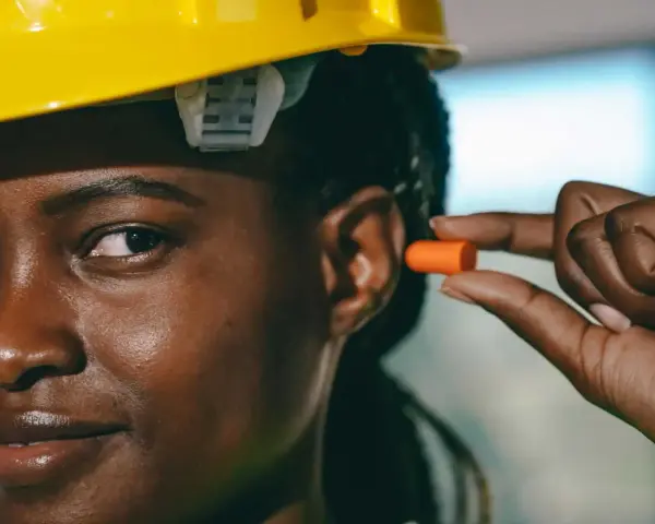 Une femme porte un casque jaune et s'apprête à insérer un bouchon d'oreille orange pour éviter le sifflement.