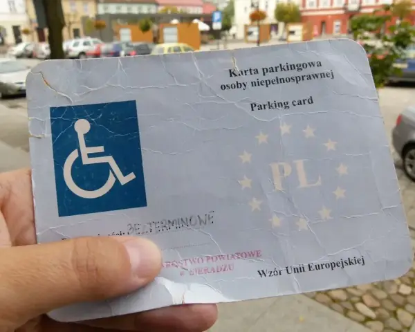 Karta parkingowa dla osoby z niepełnosprawnością, stopień umiarkowany. W tle samochody i budynki.