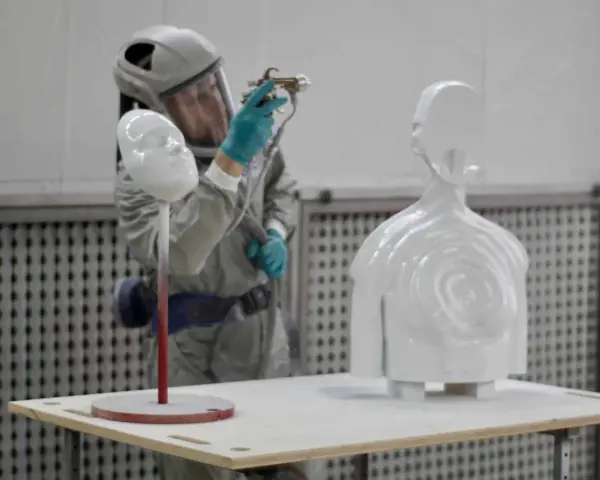 A person in a protective suit sprays a white 3D printed bust and torso, demonstrating how to paint 3d prints for a smooth finish.