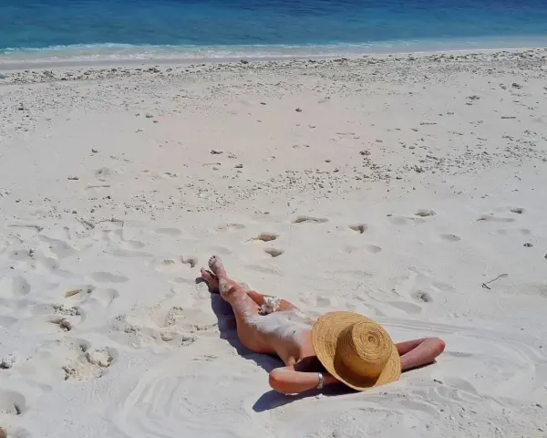 Słoneczna plaża, błękitne morze i osoba opalająca się na piasku. W tle widać fale i biały piasek z licznymi śladami stóp.