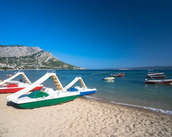 Plaża z łódkami i zjeżdżalniami, idealna na wakacje w camping Galeb. W tle góry i turkusowe morze.