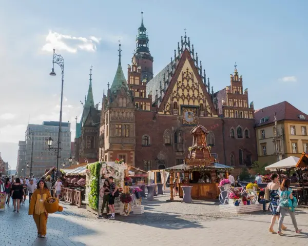 Tłum ludzi spaceruje po starym rynku we Wrocławiu, podziwiając piękną architekturę i letnie stoiska.
