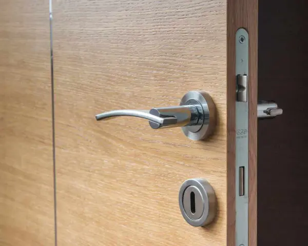 Maniglia e serratura di una porta in legno chiaro, con un'ombra scura che suggerisce una porta che cigola.