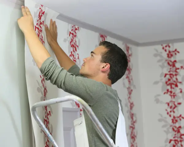 Mann tapeziert mit Vliestapete eine Wand mit rotem Blumenmuster.