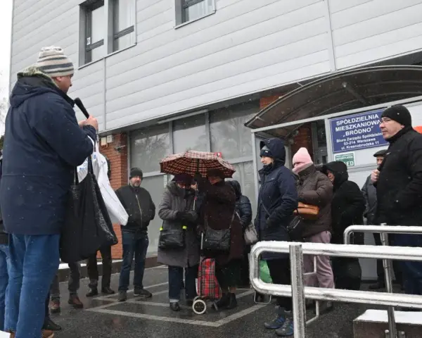 Grupa ludzi stoi przed budynkiem spółdzielni mieszkaniowej w deszczu, niektórzy pod parasolem.