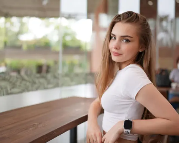 Uśmiechnięta blondynka w białej bluzce, z zegarkiem na ręku, siedzi przy stoliku. Czy to jej sekret, jak uwieść faceta?