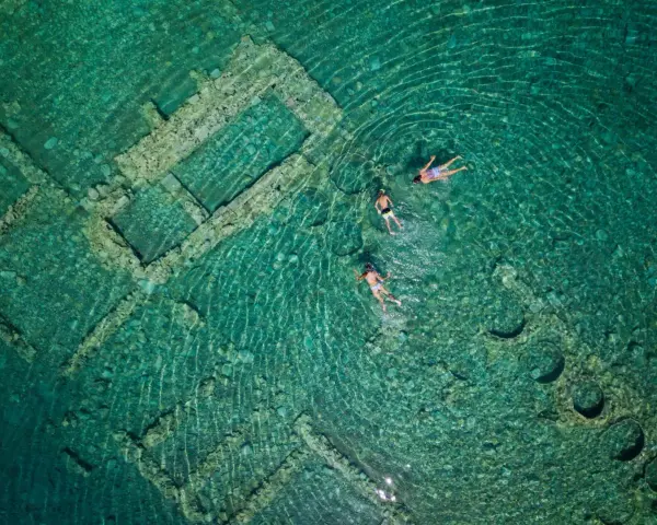 Swimmers explore the sunken city of Epidaurus, its ancient stone structures visible beneath the clear turquoise water.