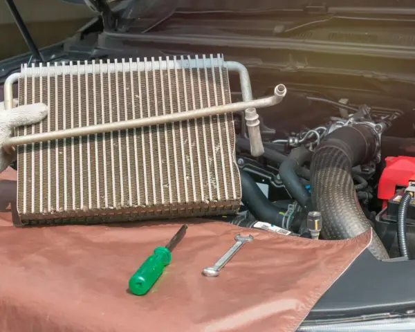 Gloved hand holds car AC evaporator. Tools and engine parts visible. Learn how to make AC colder in car by fixing this component.