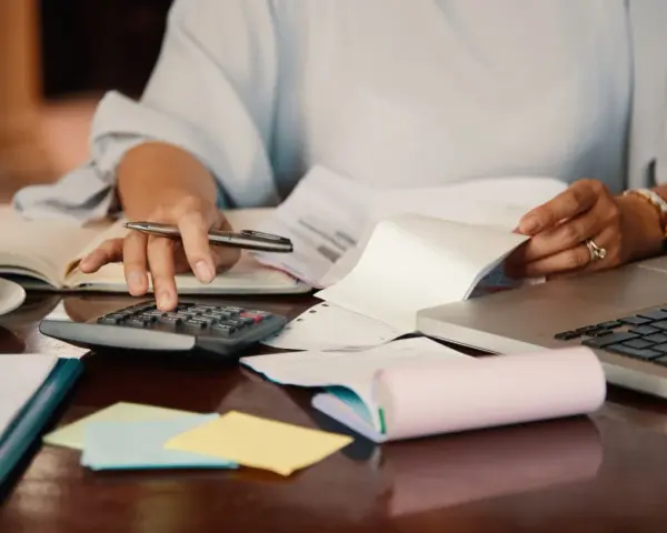 Persona revisa documentos y usa calculadora para el modelo 184. Hay café, portátil y notas adhesivas.