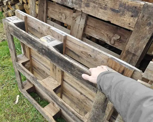 Maintenant, la question se pose : peut-on brûler du bois de palette dans une cheminée ? Une main touche des palettes en bois usagées, empilées sur de l'herbe.