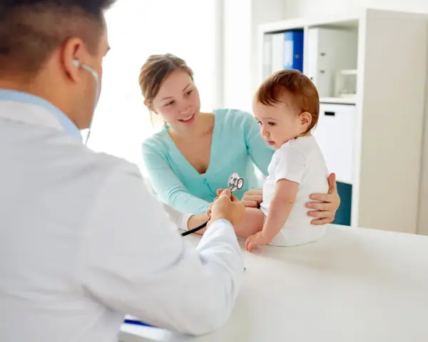 Pediatra bada niemowlę podczas pierwszej wizyty. Mama z uśmiechem obserwuje badanie.
