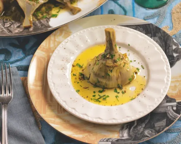 Un carciofo alla romana, tenero e profumato, adagiato su un piatto bianco con olio dorato e prezzemolo tritato.