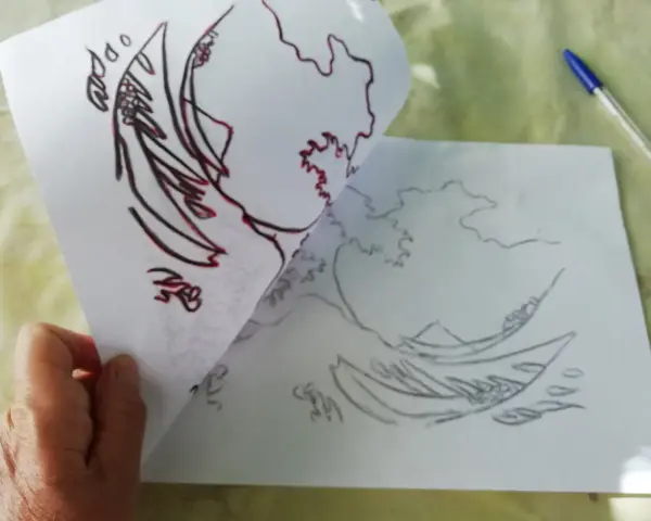 Une main tient une feuille avec un dessin de vague, prête à comment décalquer sur une toile. Un stylo bleu est visible.