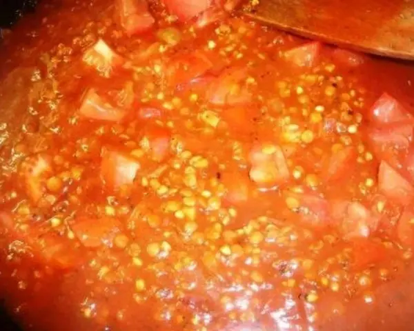 Buchweizen kochen mit Tomaten. Ein Holzlöffel rührt in der roten Sauce mit den Buchweizenkörnern und Tomatenstücken.