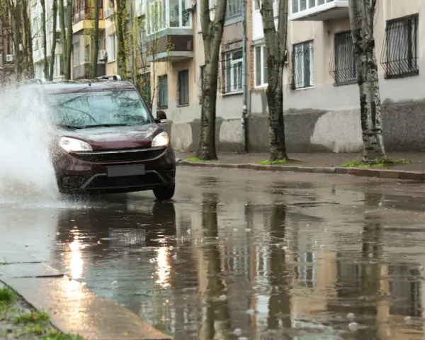 Ciemny samochód wpada w kałużę, powodując potężny **aquaplaning**. Woda rozpryskuje się na boki, a odbicia budynków i drzew tańczą na mokrej nawierzchni.