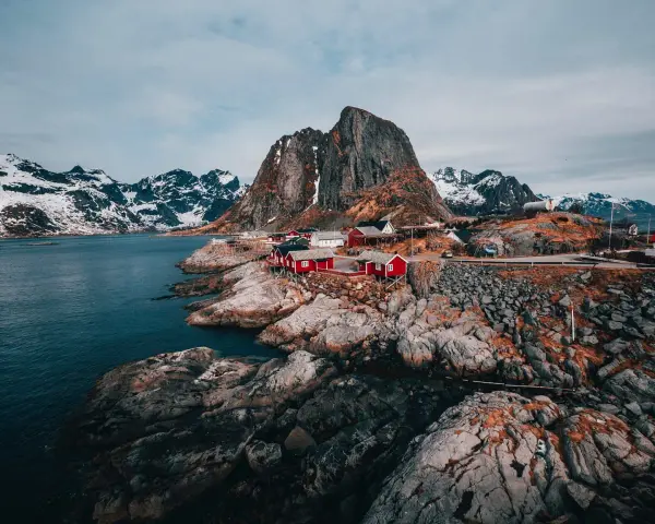 Röda fiskestugor vid en klippig kust med snöklädda berg i bakgrunden. En av de fina platserna i Norge.