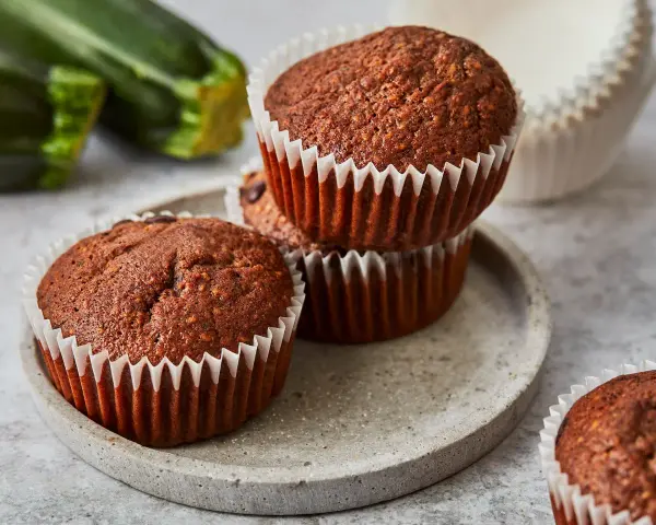 Saftige zucchini schoko muffins auf einem Steinplattenteller, im Hintergrund frische Zucchini.