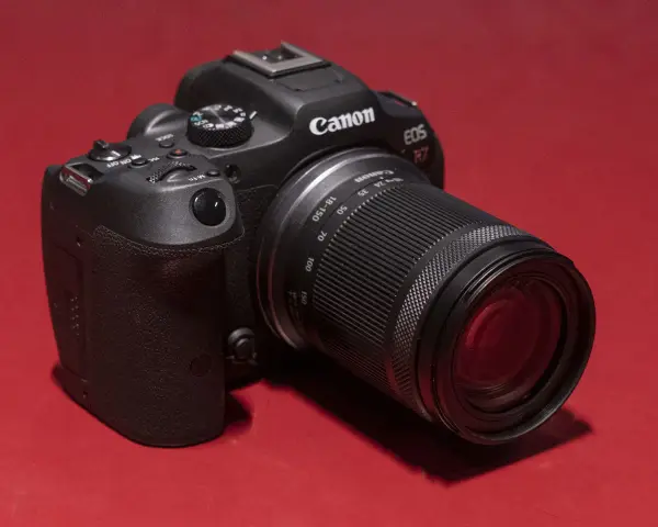 Appareil photo noir Canon EOS R7 avec objectif 18-150 mm, posé sur un fond rouge vif.