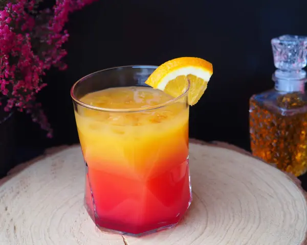 Kolorowy drink z koniakiem, ozdobiony plasterkiem pomarańczy, na tle karafki z bursztynowym alkoholem i kwitnących wrzosów.