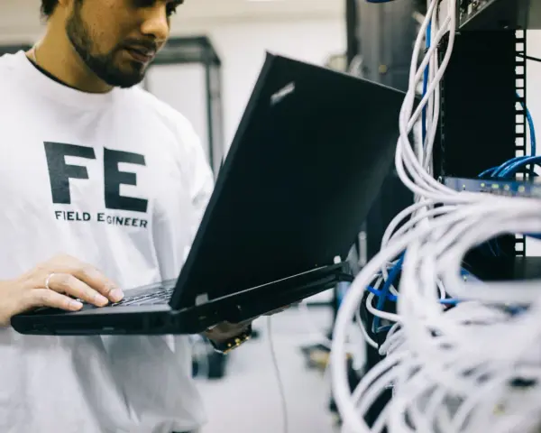 Inżynier w koszulce "FE FIELD ENGINEER" używa laptopa do sprawdzenia **trace route cmd** w serwerowni z plątaniną kabli.