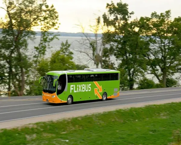 Zielony autobus Flixbus jedzie drogą przy jeziorze otoczonym drzewami.