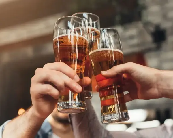 Trzy ręce unoszą kieliszki ze złotym piwem, tworząc toast. Każde piwo w szklance wygląda apetycznie.