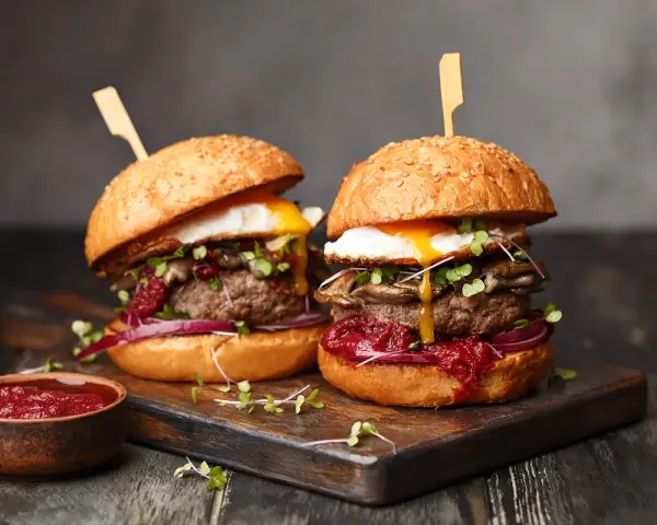 Zwei saftige Burger mit Spiegelei, Pilzen und roten Zwiebeln, serviert auf einem Holzbrett. Ein Muss für jeden Burger Laden in Berlin.