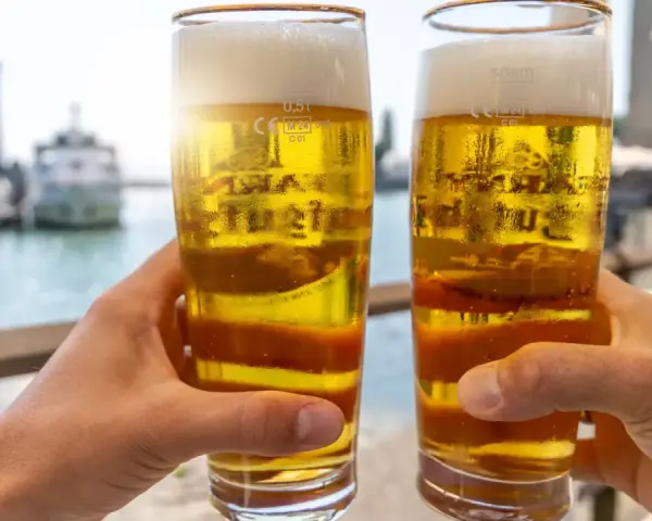 Zwei Hände halten Biergläser mit schaumigem Bier. Im Hintergrund ein Hafen. Ist Bier gesund? Ein Genuss am Wasser.