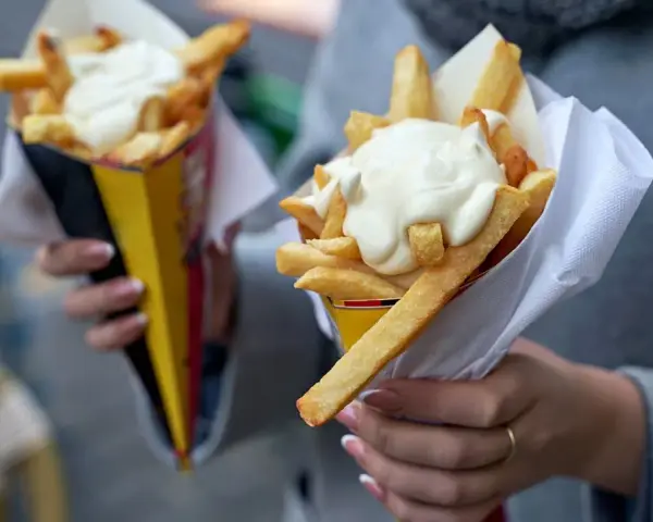 Dwie porcje frytek z majonezem w papierowych opakowaniach, trzymane w dłoniach.