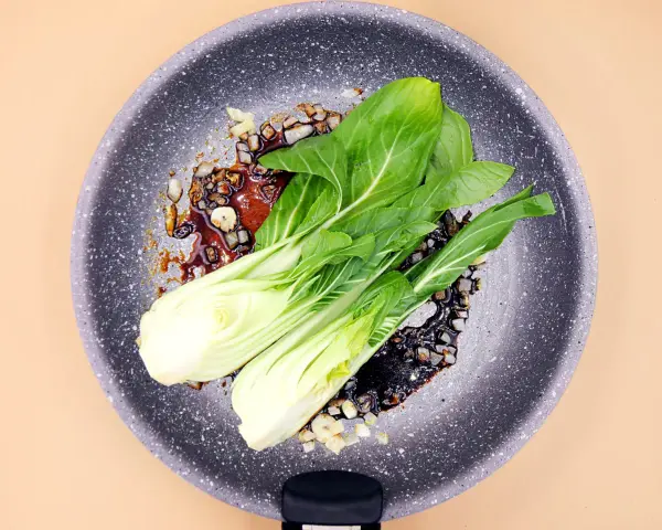 Na patelni smaży się aromatyczny sos z czosnkiem i cebulą, a na nim leży świeża sałata pak choi, gotowa do przygotowania pysznego dania.