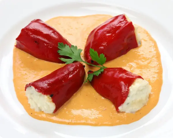 Cuatro pimientos rojos rellenos de bacalao, servidos sobre una cremosa salsa naranja y decorados con perejil.