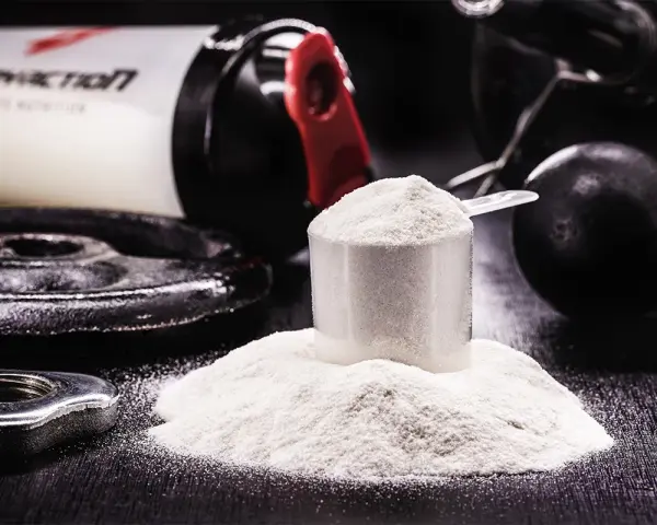 Polvo blanco en una cuchara medidora, junto a pesas y un bote de suplemento. Descubre para qué sirve la creatina y potencia tus entrenamientos.