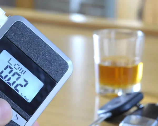 Alkomat z wyświetlaczem pokazującym 0.02% BAC, obok kieliszek z alkoholem i kluczyki samochodowe.