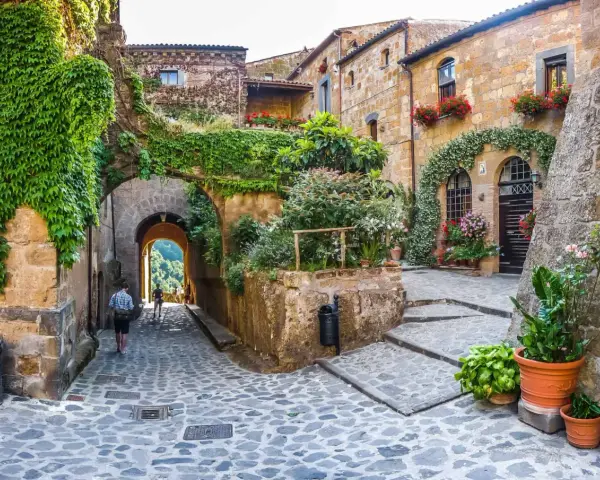 Un vicolo acciottolato con case in pietra ricoperte di edera e fiori. Un uomo cammina verso un arco che si apre su un paesaggio verde. Uno dei tanti posti belli nelle Marche.