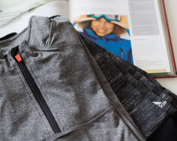 Szara bluza sportowa z zamkiem i pomarańczowym suwakiem, obok otwartej książki o narciarstwie. Czy poliamid to dobry materiał na odzież sportową?